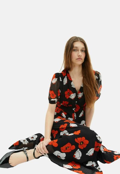 Junge Frau mit langen Haaren, die sitzt und ein schwarzes Kleid mit rotem und weißem Blumenmuster sowie schwarze Schuhe mit Knöchelriemen trägt, vor einem weißen Hintergrund.