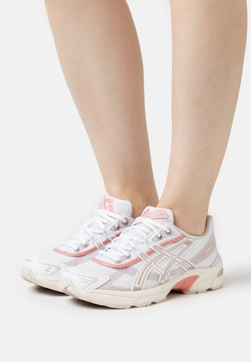 Fødder iført hvide og lyserøde ASICS løbesko med snørebånd, stående på en ensfarvet hvid baggrund.