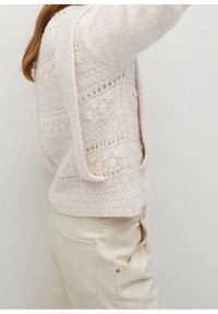 Pull en tricot rose pâle, léger, avec des motifs texturés et des côtés ouverts, associé à un pantalon beige taille haute avec des poches latérales.