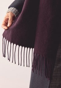 Dziļi purpura šalle no mīksta auduma ar bārkstīm malās un gludu tekstūru, pārklāta pār pelēku apģērbu ar rakstainu detaļu.