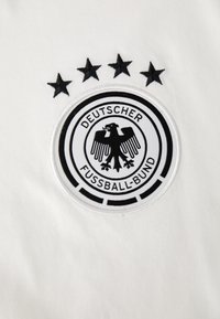 Hvit tekstil med et sirkulært svart brodert logo med en ørn og teksten "DEUTSCHER FUSSBALL-BUND," over fire svarte stjerner.