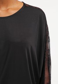 Czarna bluzka z okrągłym dekoltem, długimi przezroczystymi koronkowymi rękawami oraz gładkim, lekkim materiałem o lekko luźnym kroju.