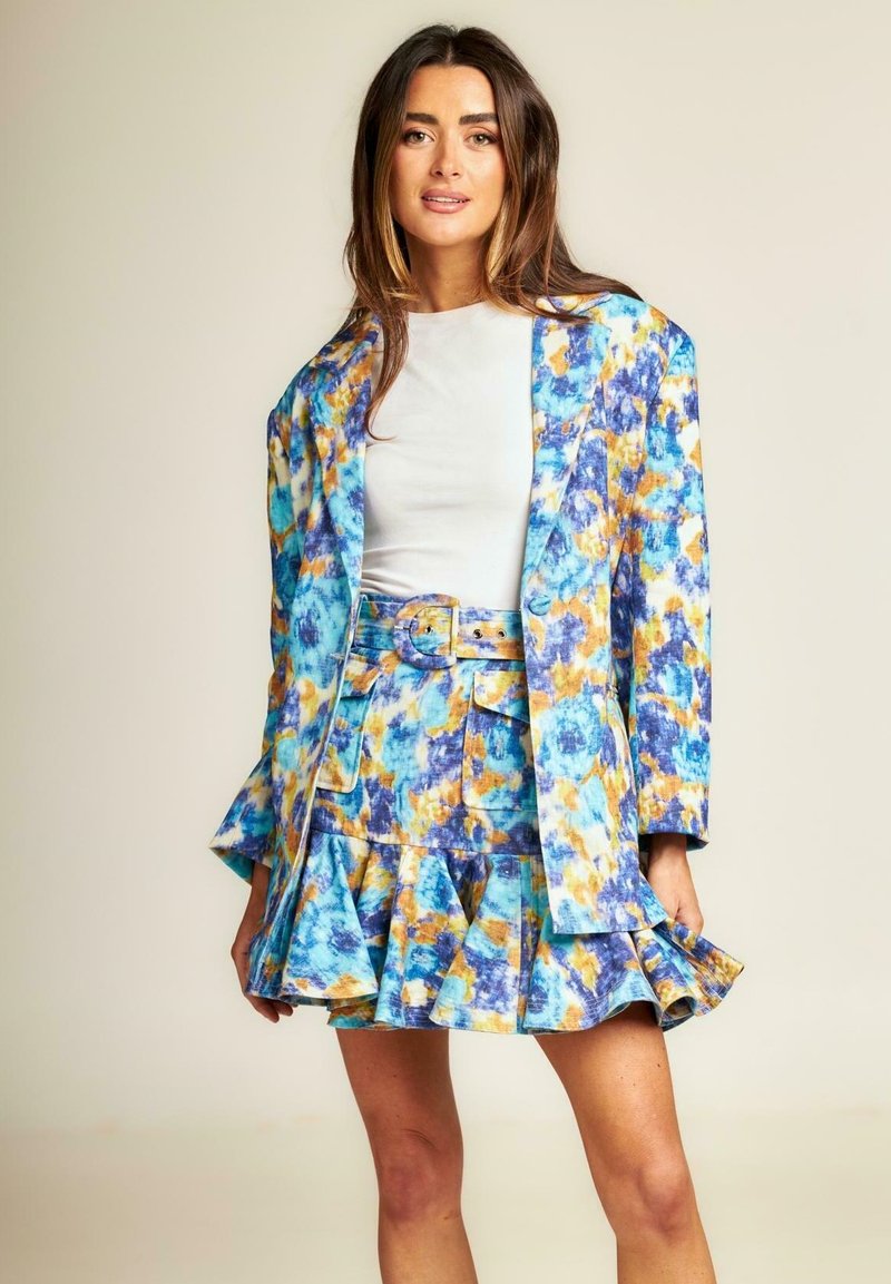 Blazer con fantasia floreale blu, gialla e bianca abbinato a una gonna plissettata. Presenta una cintura e maniche corte con vestibilità rilassata.