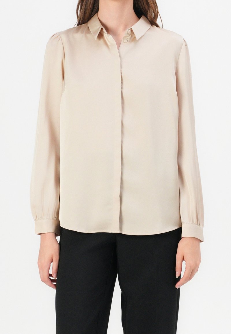 Femme portant un chemisier beige à manches longues boutonné avec col et un pantalon noir, debout devant un fond blanc uni.