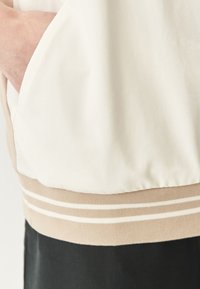 Chaqueta bomber blanca con puños acanalados en beige, que presenta una textura lisa y mate y un bolsillo lateral visible a lo largo del dobladillo.