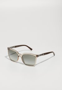 Ochelari de soare cu un cadru transparent bej și lentile gri cu degradé. Brațele maro au un model țesut și logo-ul "vogue" pe lateral.