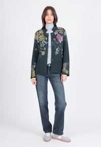 Cardigan floreale verde scuro con accenti gialli e rosa, chiusura con bottoni e tasche frontali, abbinato a un dolcevita grigio e jeans blu.
