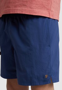 Costumi da bagno blu navy con una texture leggera, caratterizzati da uno spacco laterale e un accento arancione con la lettera "F" vicino all'orlo.