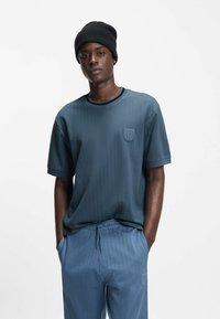 Siniraidallinen t-paita, jossa on resorikaulus ja paikkatasku. Yhdistetty samaan sarjaan kuuluvien nyörillä kiristettävien housujen kanssa. Musta neulottu pipo päässä.