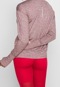 Maglia sportiva a maniche lunghe color marrone scuro con fori per i pollici, abbinata a leggings aderenti di un rosso brillante con pannelli testurizzati e dettagli in rete.