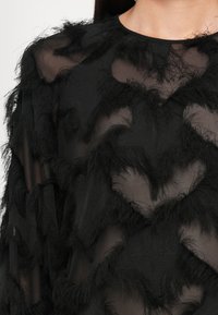 Top nero trasparente con un design testurizzato e accenti frangiati e ondulati. Il materiale sembra leggero ed è stratificato per una profondità visiva.