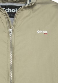 Beige Jacke mit einem Reißverschluss vorne, ausgestattet mit einem gestickten "Schott"-Logo in Weiß und Rot. Strukturierter Stoff mit einer glatten, matten Oberfläche.