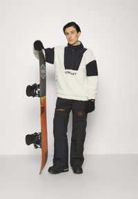 Weißer und schwarzer Fleece-Pullover mit hohem Kragen, kombiniert mit schwarzen Schneehosen und Handschuhen. Person hält ein Snowboard mit orangefarbenen Akzenten.