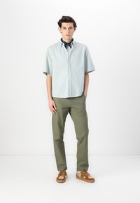 Chemise bleu clair à manches courtes avec boutons, pantalon vert olive, chaussures brunes à lacets et une écharpe bleue nouée autour du cou. Fond uni.
