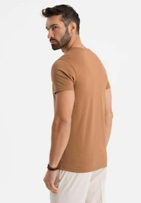 T-shirt in cotone marrone con vestibilità classica, scollo rotondo, maniche corte e texture liscia, indossato da una persona con pantaloni chiari.