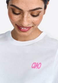 White cotton t-shirt featuring pink embroidery of the word "CIAO." Simple round neckline and smooth texture. Close-up view of the shirt.