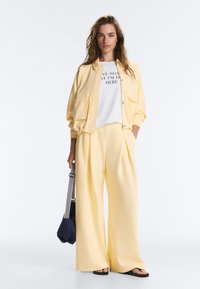 Jeune femme porte un pantalon jaune à jambes larges, une veste assortie, un t-shirt blanc à motif graphique, des sandales noires et tient un sac bleu marine avec des bretelles rayées.