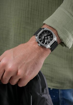 Schwarze Uhr mit Edelstahlgehäuse, strukturiertem schwarzen Armband, markanten weißen Ziffern und schwarzem Ziffernblatt. Wohl am Handgelenk, das eine schwarze Jacke hält.