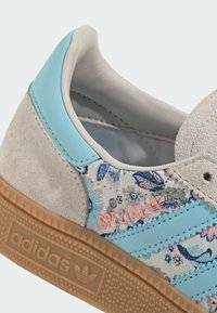 Lähikuva Adidas Spezial -tossusta, jossa on siniset nahkaiset raidat, kukkakuvioinen kangas, mokkanahkainen kanta ja kumipohja, jossa on Adidas-logo.