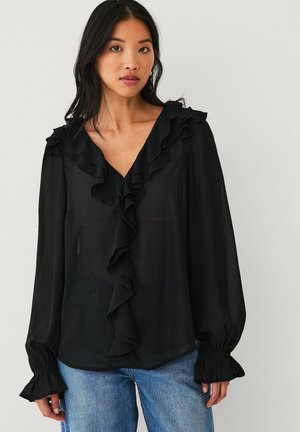 Kobieta w przezroczystej czarnej bluzce z falbanami przy dekolcie w kształcie litery V i mankietach, w połączeniu z niebieskimi jeansami, stoi na tle gładkiego jasnego tła.