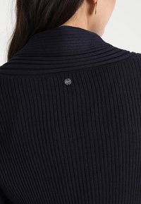 Frau trägt einen schwarzen gerippten Pullover, zeigt den Rücken mit einem kleinen runden Metallschild in der Nähe des Kragens und einem goldenen Creolenohrring.