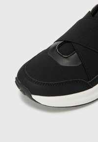 Scarpe sportive nere con tomaia strutturata, dotate di una fascia elastica sulla parte anteriore, dettagli lucidi e suola bianca.