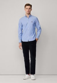 Camisa de botones a rayas azules y blancas, con cuello y logotipo, combinada con pantalones oscuros y zapatillas blancas. Textura de tela suave.