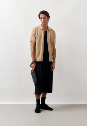Jeune homme portant une chemise beige à manches courtes boutonnée, un pantalon court noir, des chaussures et des chaussettes noires, tenant une pochette noire sur un fond uni.