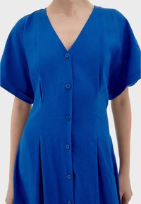 Abito in lino blu con scollo a V, maniche corte e chiusura frontale con bottoni. Presenta una silhouette aderente e una consistenza morbida.