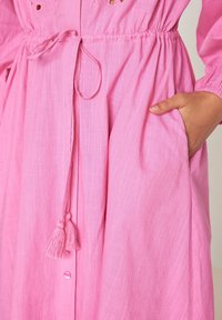 Robe en lin rose dotée d'une taille élastique avec une ceinture à nouer, de poches et d'une fermeture à boutons avec un détail de pompon. Tissu texturé avec des découpes en forme de cœur.