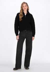 Maglione nero lavorato a maglia con zip e colletto alto, caratterizzato da un motivo testurizzato, abbinato a jeans scuri a vestibilità larga e scarpe nere.
