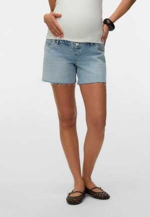 Kvinde iført lyseblå cowboyshorts, hvid top, sort armbånd og sorte flettede flade sko, stående med hænderne nær hofterne.