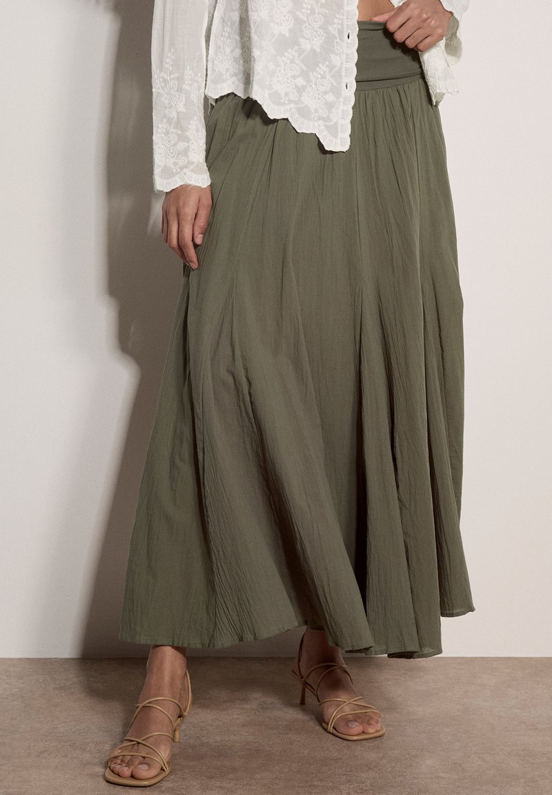 Femme portant un haut en dentelle blanche, une longue jupe vert olive et des sandales à talons beiges à brides, debout devant un fond uni.