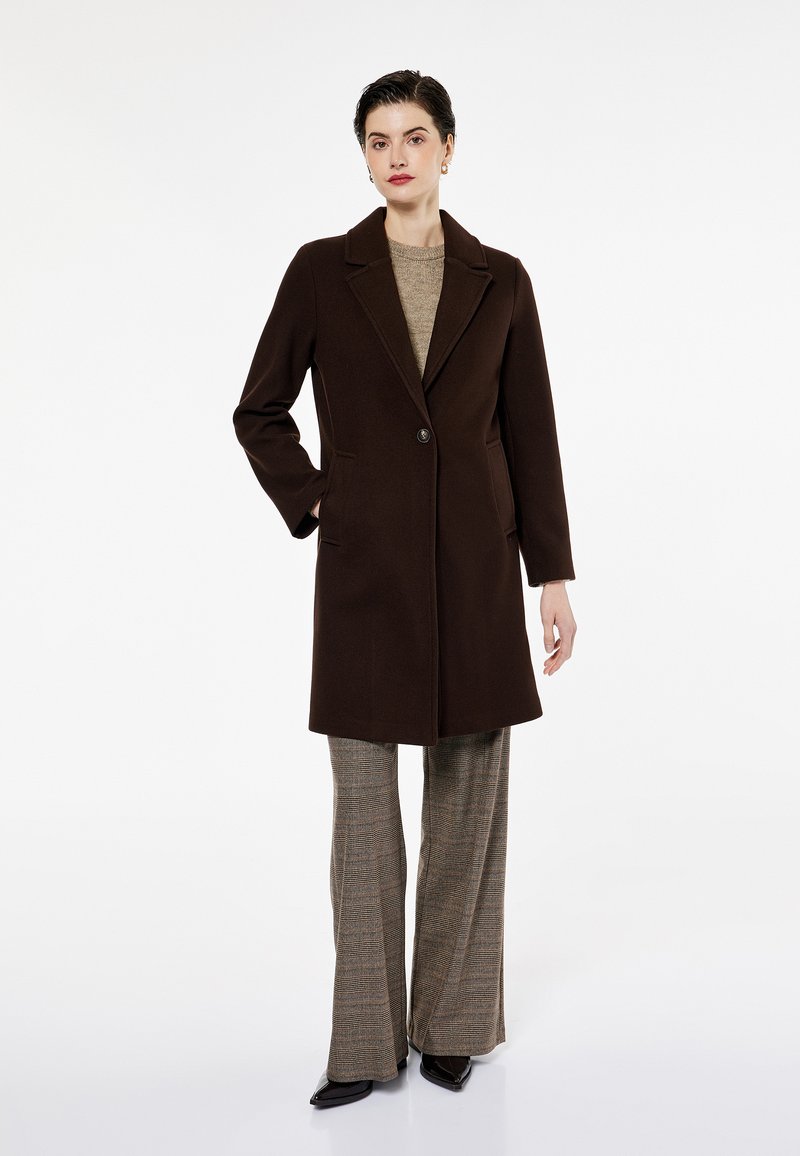 Ruskea villatakki, jossa on viistot kaulukset, yksittäinen nappilukitus ja kaksi sivutaskua. Yhdistetty ruudullisiin leveälahkeisiin housuihin.