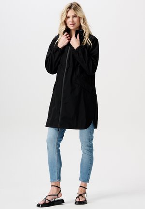 Schwarze wasserdichte Jacke mit hohem Kragen, Frontreißverschluss und Seitentaschen, kombiniert mit hellblauen Skinny-Jeans und schwarzen Riemchensandalen.