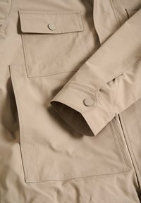 Jachetă bej cu o textură netedă, având un buzunar mare pe piept și o manșetă cu nasture. Design simplu, minimalist, cu linii clare.