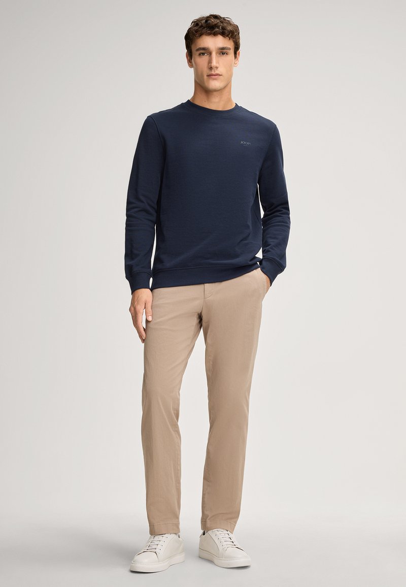Maglietta a maniche lunghe blu navy con collo a giro, polsini a coste, abbinata a pantaloni beige e sneakers bianche. La maglietta presenta un logo discreto.