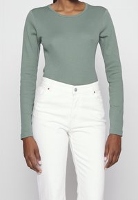 Pitkähihaisen, istuvan yläosaan pehmeästä vihreästä kankaasta, jossa on pyöreä pääntie; yhdistettynä valkoisiin rakenteellisiin farkkuihin, joissa on napitus edessä.