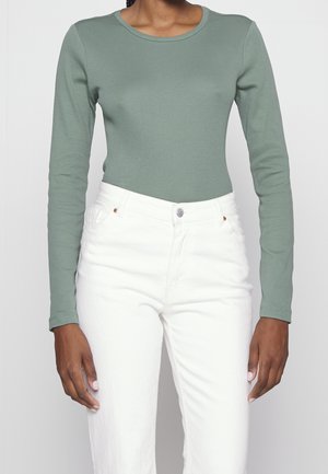 Langærmet, tætsiddende top i blødt grønt stof med rund halsudskæring; parret med hvide teksturerede jeans med knaplukning foran.