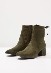 Botas de tobillo de ante verde oliva con punta afilada, tacón de bloque y cremallera lateral. Presenta una sutil textura de tela y una tira en la parte trasera.