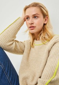 Maglione lavorato a maglia beige con texture a coste, dotato di un vivace bordo giallo lungo il colletto e le maniche, indossato con jeans blu.