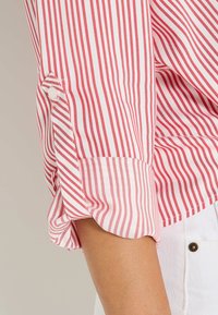 Camisa a rayas rojas y blancas con mangas remangadas, que presenta un detalle de botones y una textura de tela ligera.