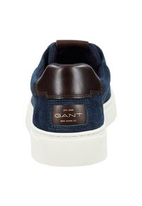 Zapatilla de ante azul marino con un tabique de cuero marrón en el talón, suela de goma blanca y detalle del logo en dorado. Parte superior texturizada con un diseño limpio y minimalista.