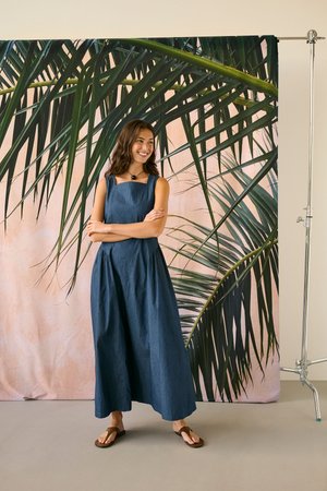 Pikkade tumedate juustega naine seisab käed risti, naeratades, seljas varrukateta sinine kleit ja pruunid sandaalid suure palmi lehepildi taustal.