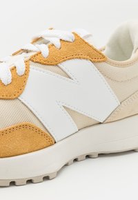 Primo piano di una sneaker beige con lacci bianchi, dettagli in suede color sabbia e grande logo "N" bianco sul lato.