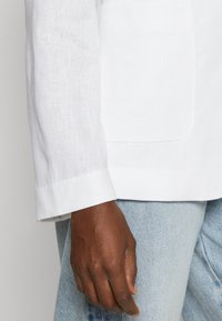 Manga de un blazer blanco de lino con un puño, que muestra un tejido texturizado y un bolsillo de parche, combinado con unos jeans de denim azul claro.