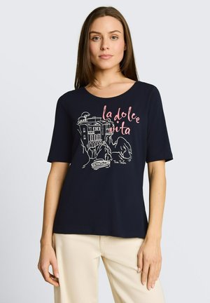 Frau trägt ein marineblaues kurzärmeliges Shirt mit „la dolce vita“ und einer Illustration eines Küstendorfs, kombiniert mit beigen Hosen, steht vor hellem Hintergrund.