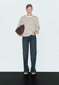 Gestreiftes Langarmoberteil in Creme und Schwarz, kombiniert mit dunklen, geraden Jeans. Hält eine braune, geschwungene Handtasche, barfuß auf einer schwarzen Oberfläche.