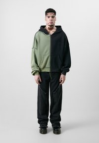 Hooded zip-up sweatshirt in olive green and black, featuring a split design and raw hems. Paired with distressed black denim pants and boots.