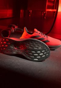 Paire de chaussures de course orange vif avec semelles et rayures noires, posées sur un sol sombre éclairé par une lumière d'ambiance rouge.